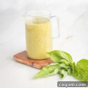 Small glass pitcher filled with mustardy yellow colored creamy dressing, with sprigs of basil in the foreground.