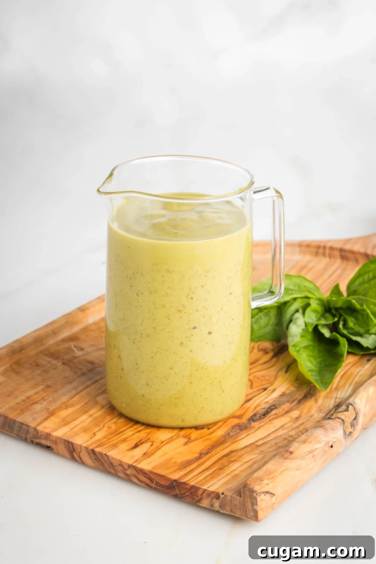 A small glass pitcher filled with a creamy, deep yellow balsamic dressing, garnished with fresh basil leaves on the side, ready to be used.