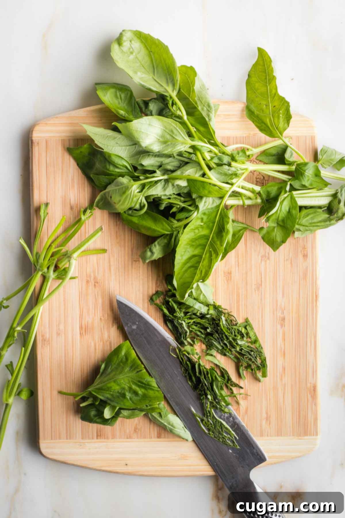 Close-up shot of fresh basil leaves, some detached from their stems, stacked neatly and being thinly sliced with a knife on a cutting board, preparing them for the salad.