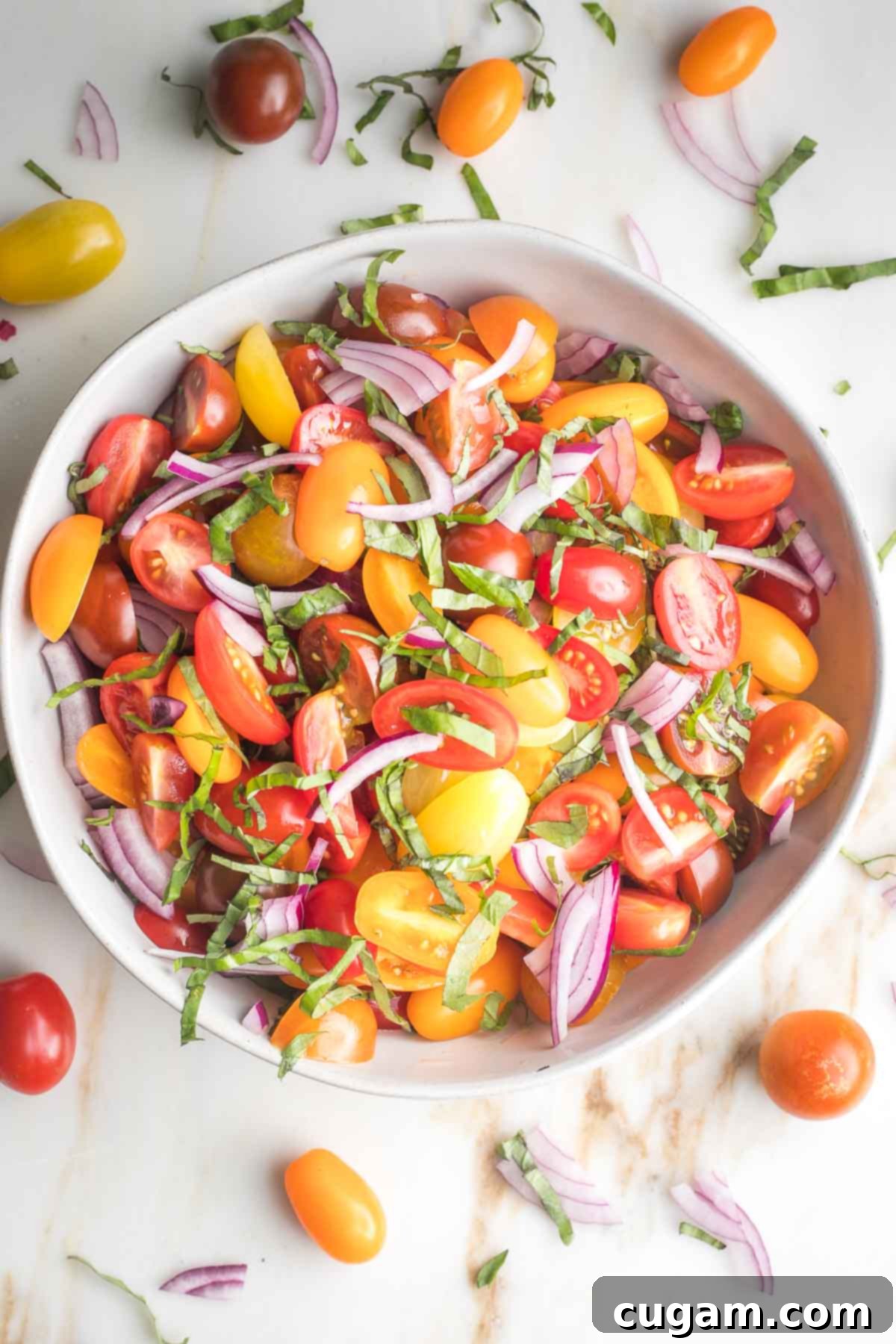 Vibrant yellow, orange, and red chopped tomatoes are mixed with delicate onion slices and fresh basil leaves in a pristine white serving bowl, showcasing the colorful medley.