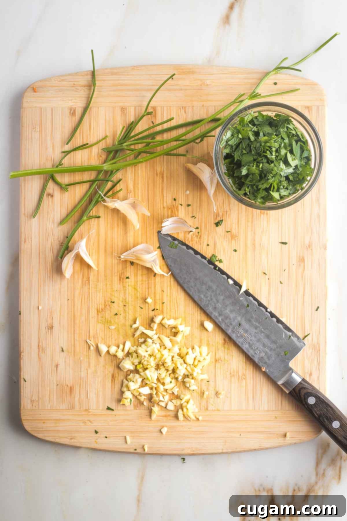 Pan-Fried Eggplant Salad 9 Chopped parsley in a small dish next to a chef's knife roughly chopping fresh garlic cloves on a cutting board.