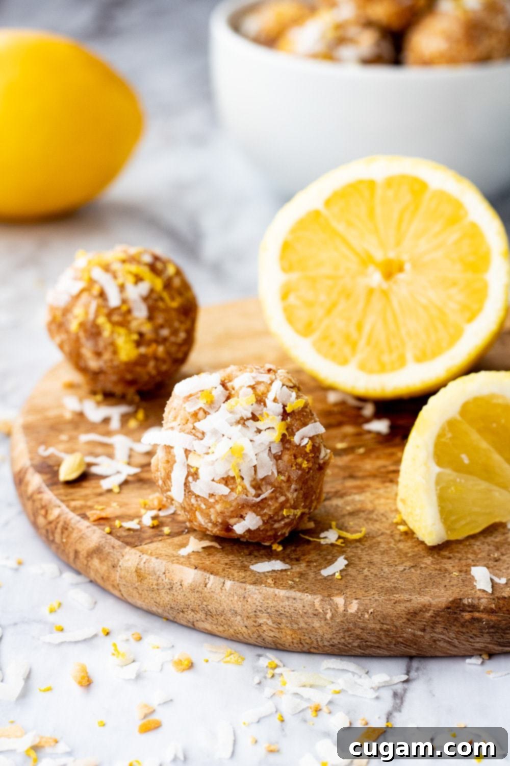 Lemon coconut protein balls are no bake energy balls with dates, oats, almond butter, almond flour, coconut, and lemon. Lemon coconut protein ball on wood board with sliced lemon on marble backdrop.