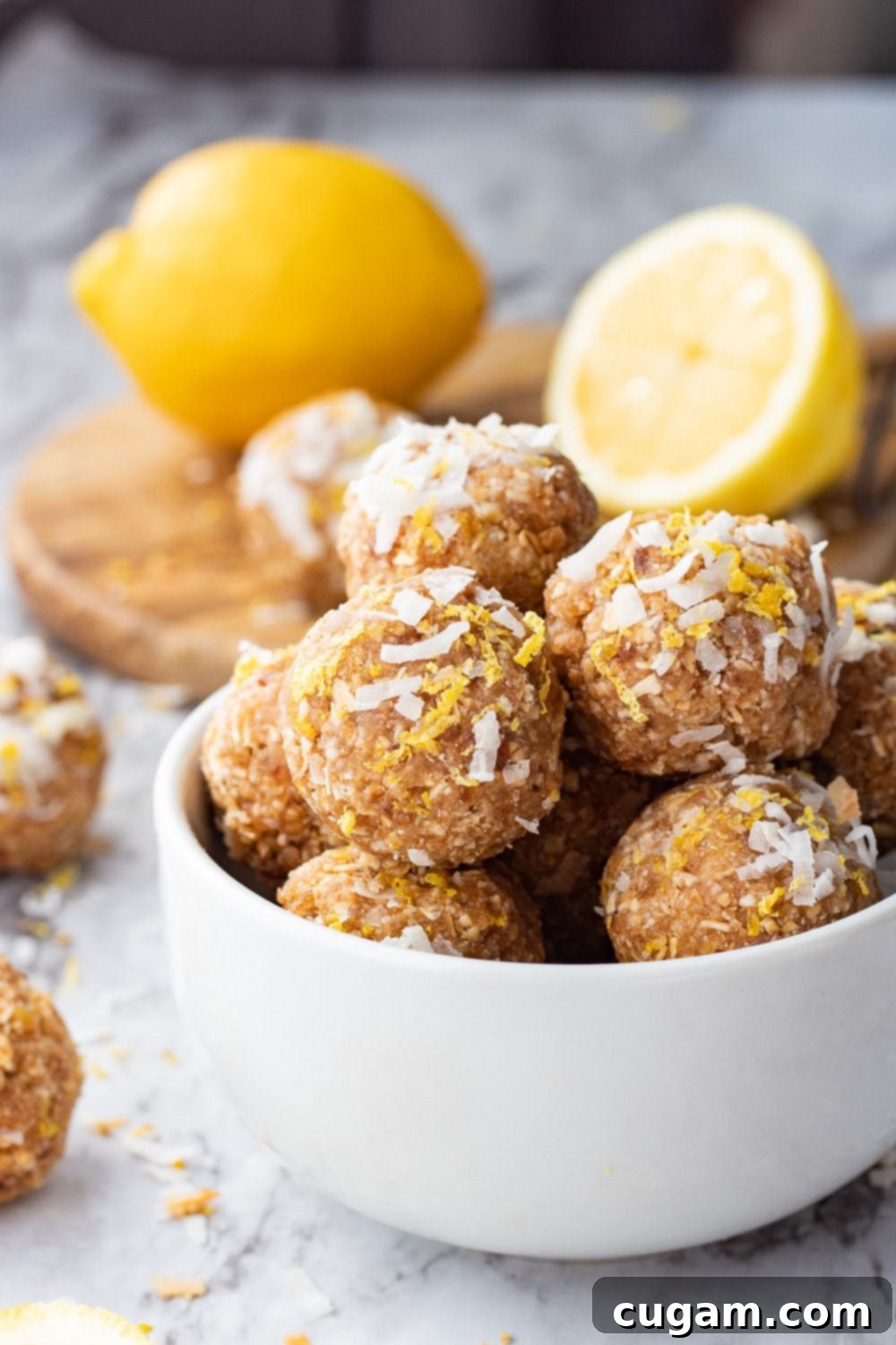 Lemon coconut protein balls are no bake energy balls with dates, oats, almond butter, almond flour, coconut, lemon, and protein powder. #proteinballs #energy bites Lemon Coconut protein balls in white bowl with whole lemon and slices in background on wood board.