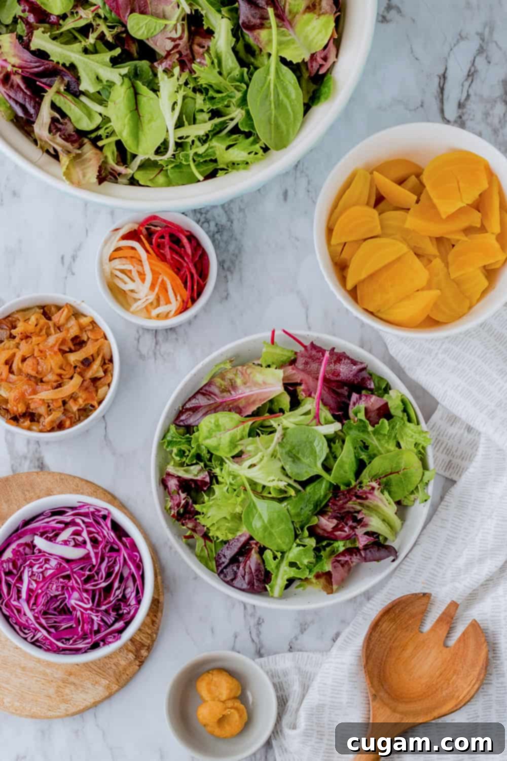 Lettuce, gold beets, caramelized onions, red cabbage, and miso in white bowls with wood spoon on marble. 