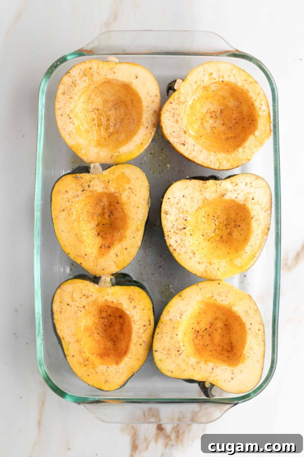 Acorn squash halves in a baking dish, cut side up, seasoned with olive oil, salt, and pepper.