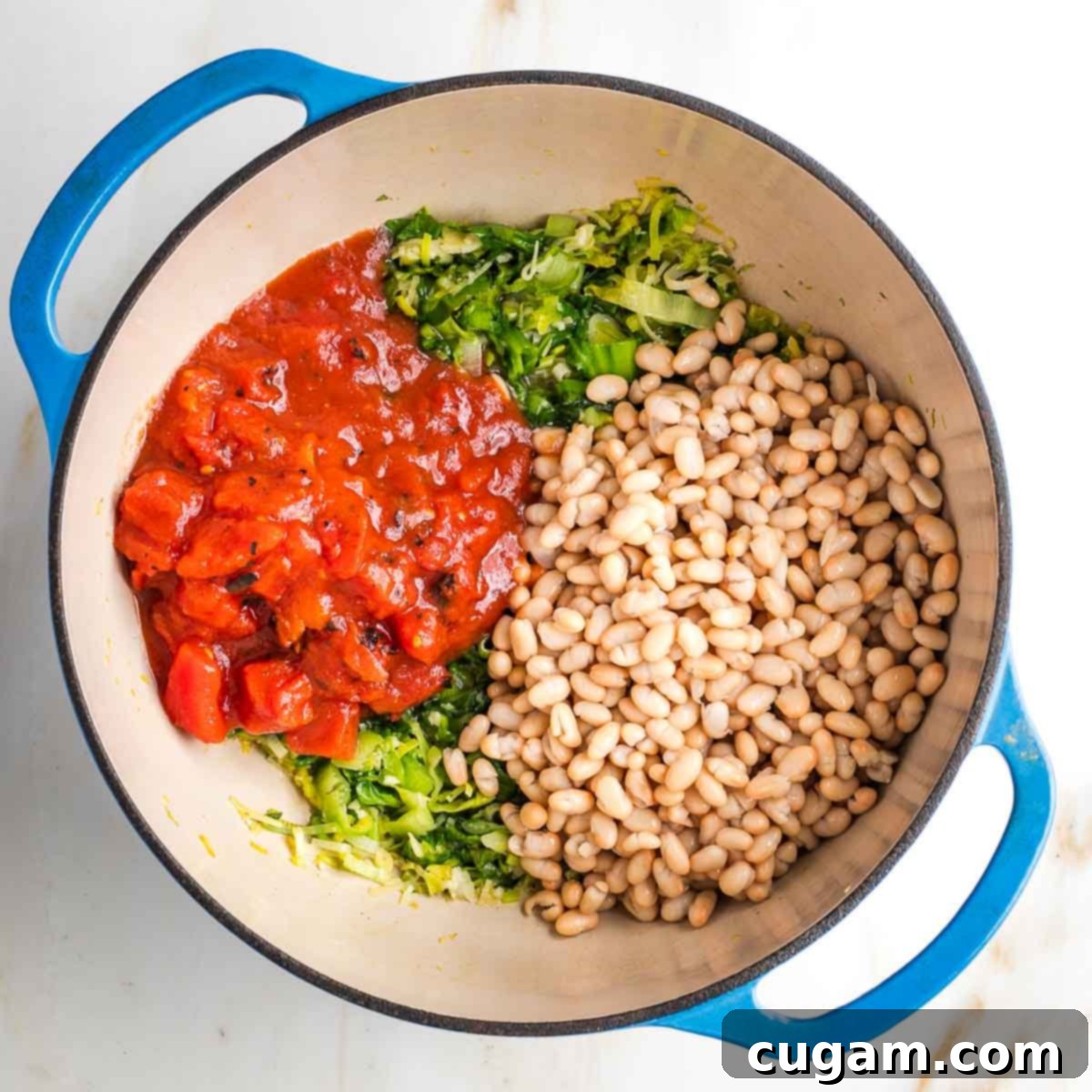 The bottom of a dutch oven filled with wilted greens, then topped with diced tomatoes and rinsed white beans, before liquid is added.