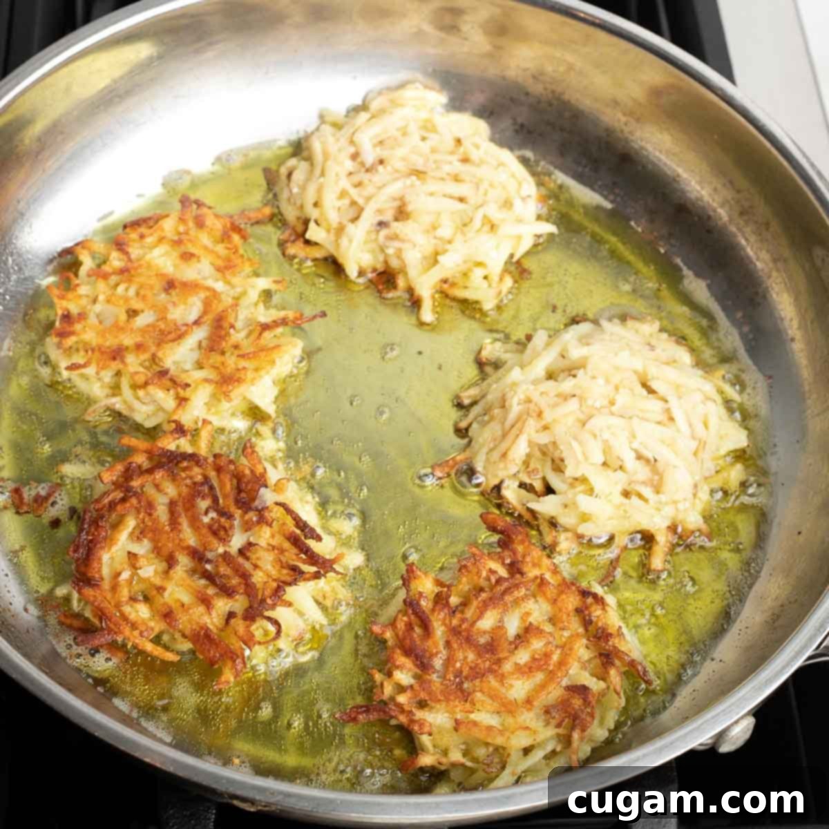 Grandma's Secret Potato Latkes 11 Potato latkes frying in a large skillet in oil, half already flipped to other side.