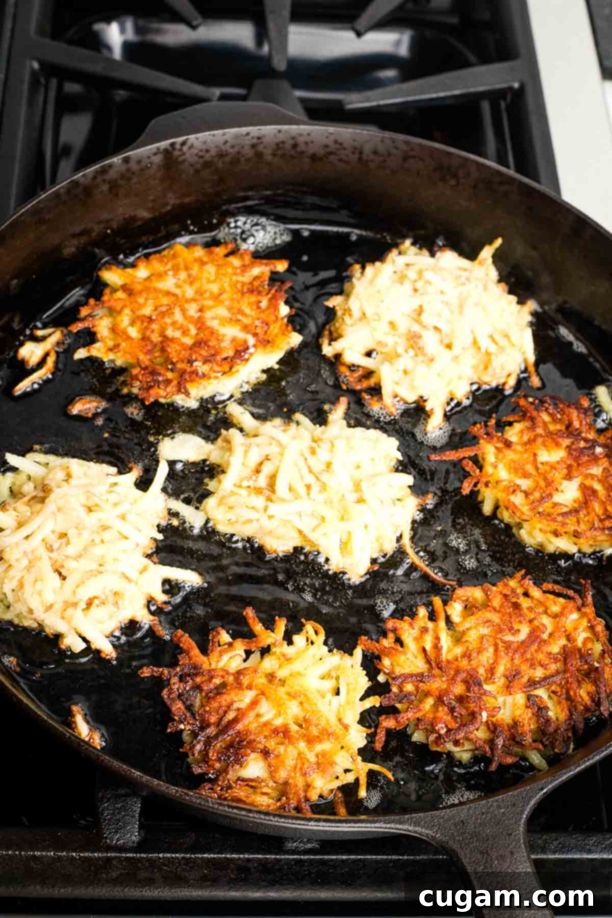 Grandma's Secret Potato Latkes 14 Cast iron skillet filled with oil and latkes, some already turned with crispy edges.