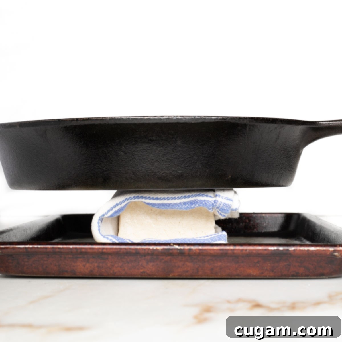 Block of tofu, wrapped in a kitchen towel with a heavy cast iron skillet on top.