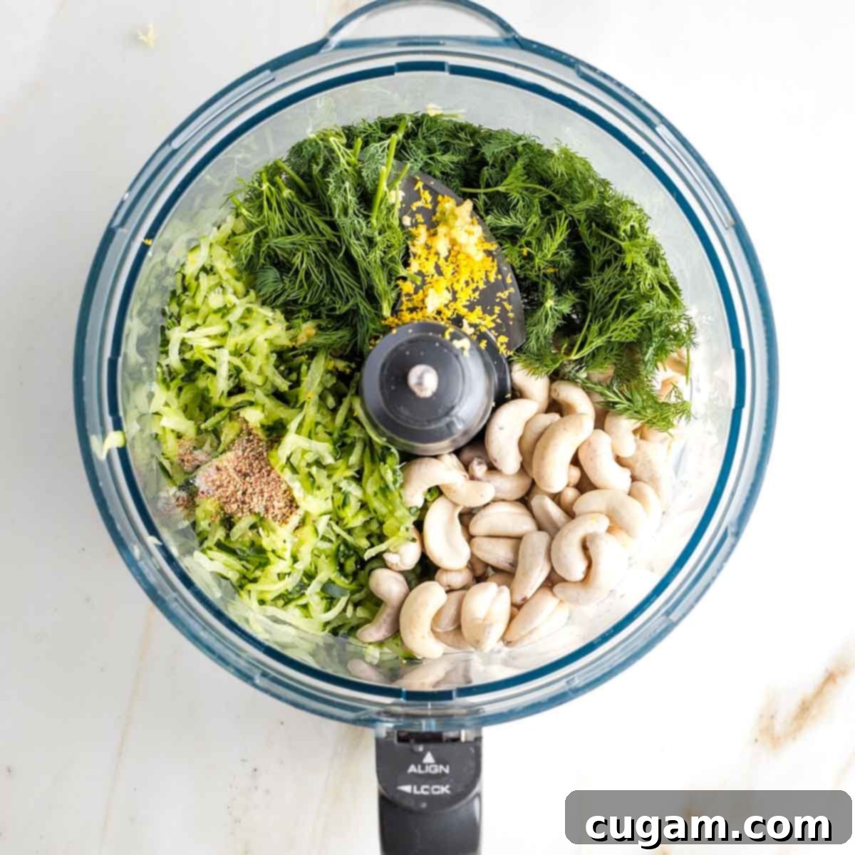 Ingredients for dairy-free tzatziki sauce in the bowl of a food processor: fresh herbs, grated cucumber, cashews, lemon zest and seasonings.
