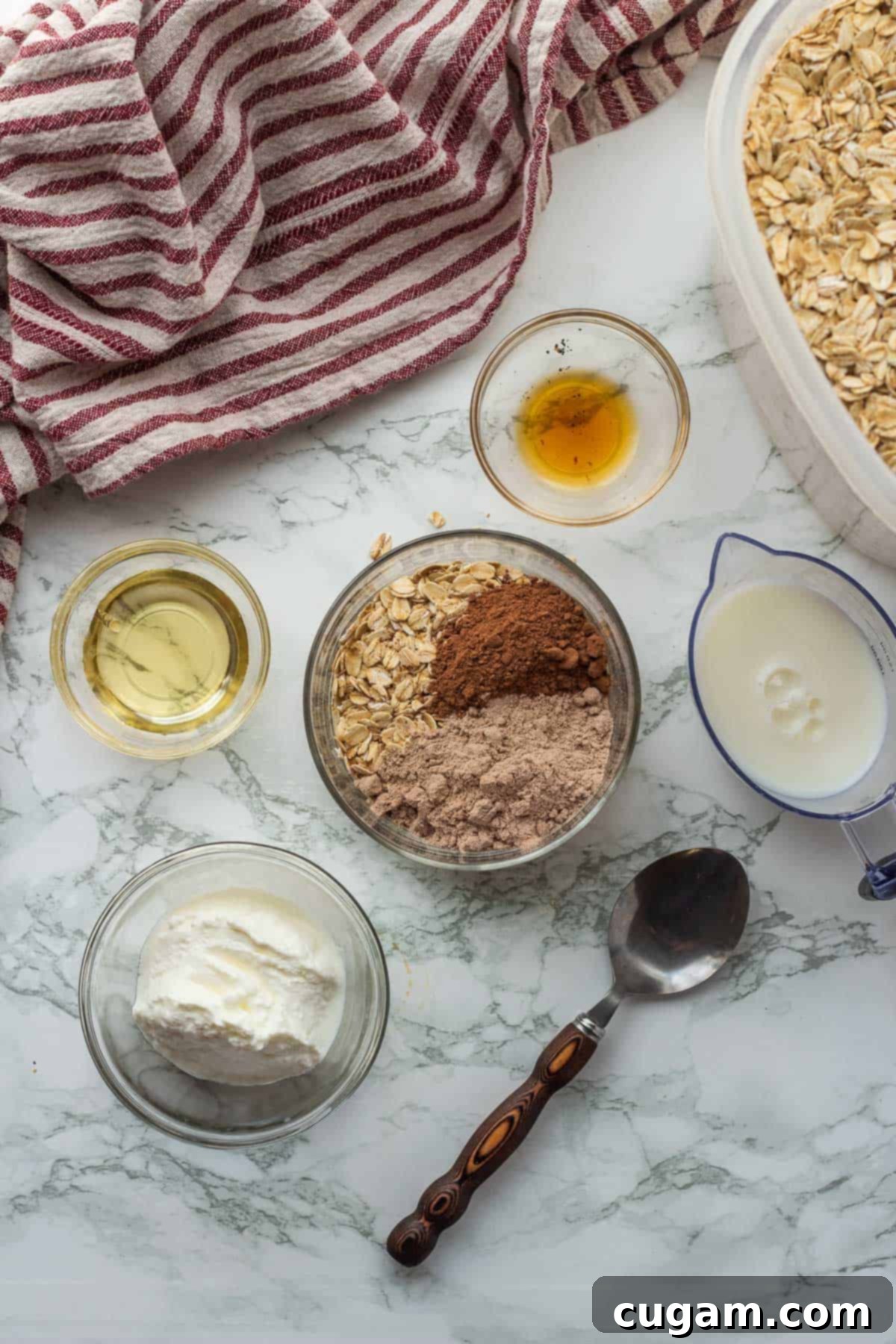 Ingredients for brownie batter overnight oats in glass bowls on marble countertop with dish towel.