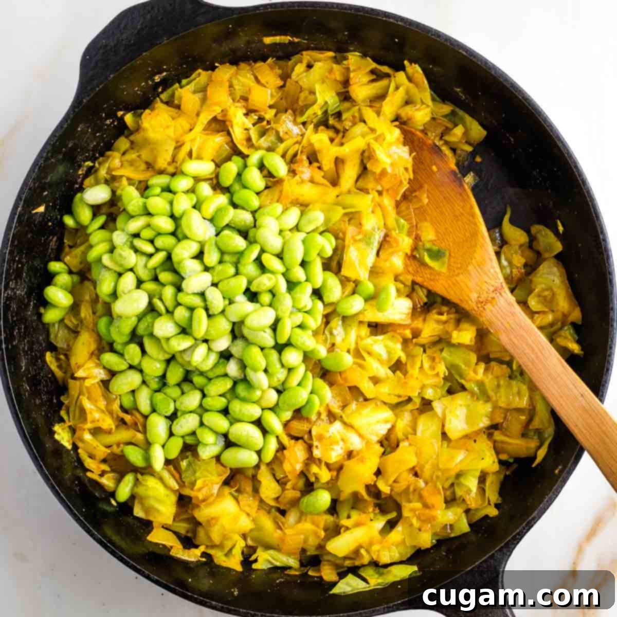 Adding shelled frozen edamame to a pan of cooked chopped cabbage.