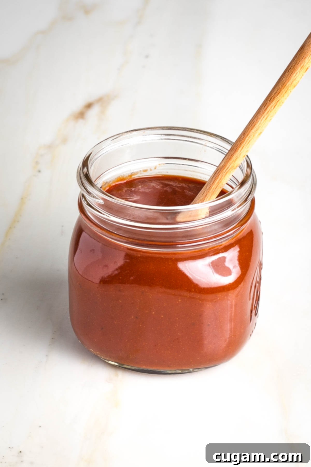 A clean glass jar filled to the brim with luscious homemade no-cook BBQ sauce, with a wooden spoon sticking out invitingly, ready for storage in the refrigerator.