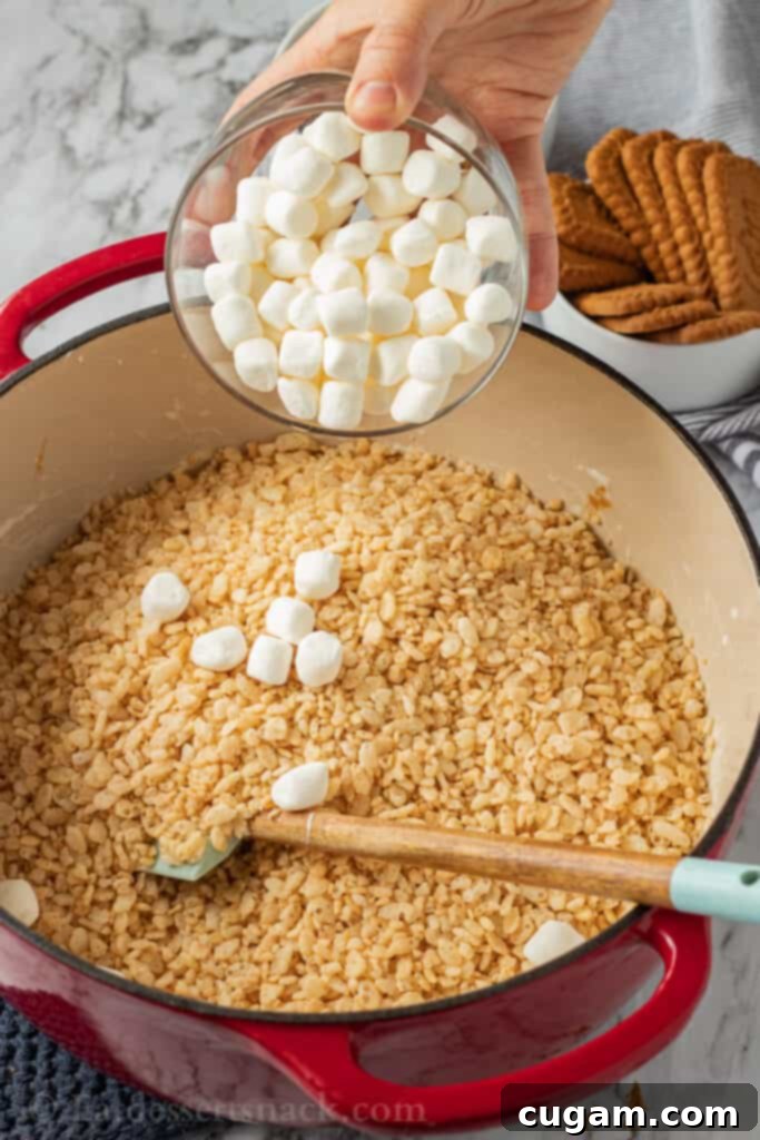 Melted marshmallows being stirred in a glass bowl over a pot, with puffed rice cereal in the background.