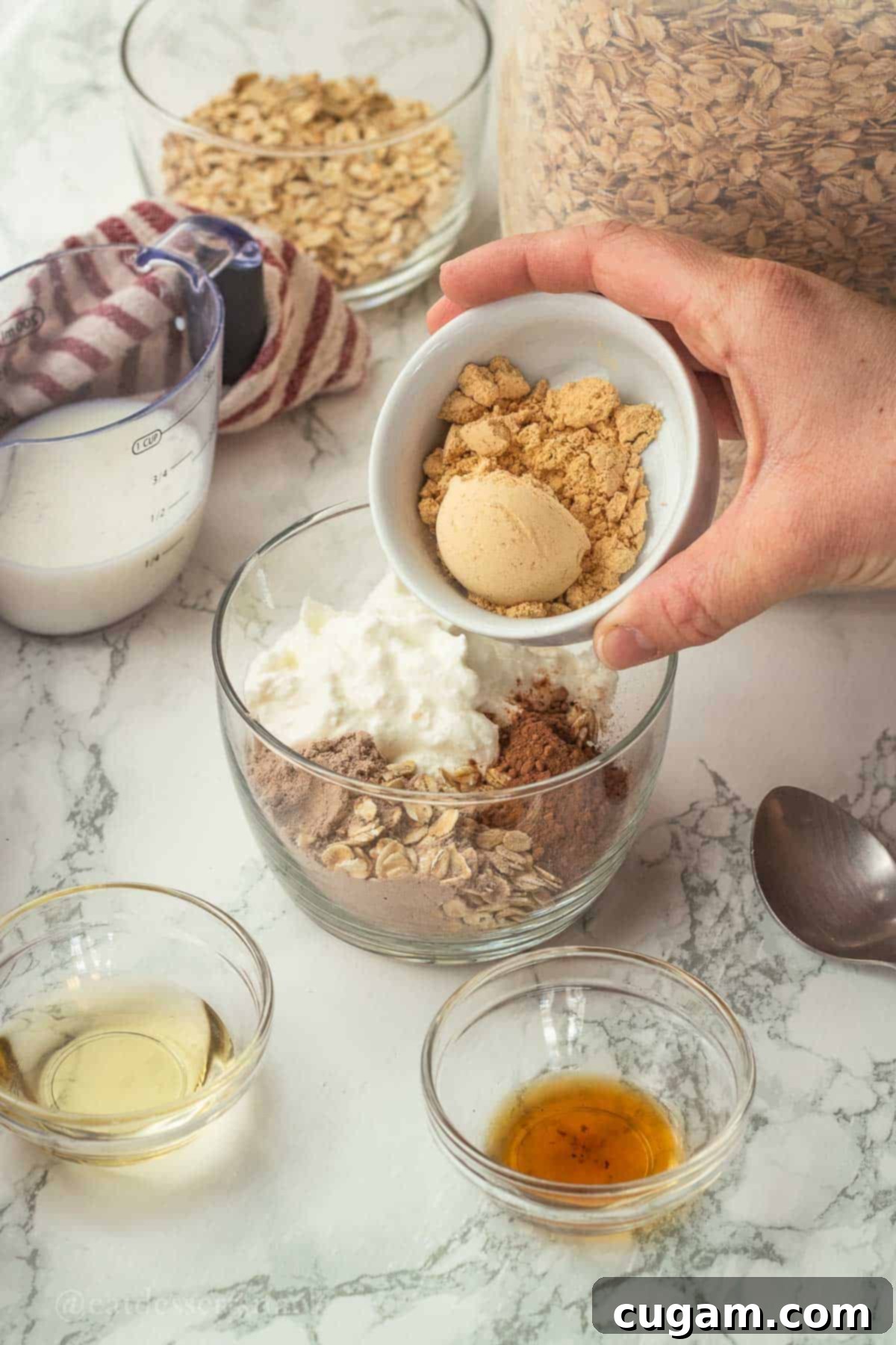 Pouring peanut butter powder into a glass bowl containing a mix of oats, Greek yogurt, cocoa powder, and chocolate protein powder, all ingredients for the chocolate peanut butter overnight oats.