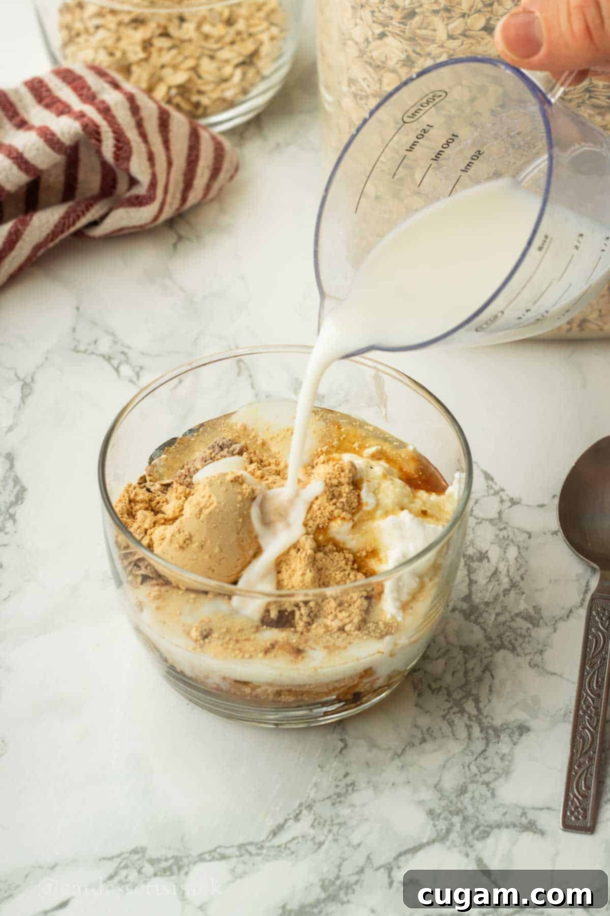Pouring milk into a glass bowl filled with the pre-mixed dry ingredients and yogurt for chocolate peanut butter overnight oats, indicating the next step in preparation.