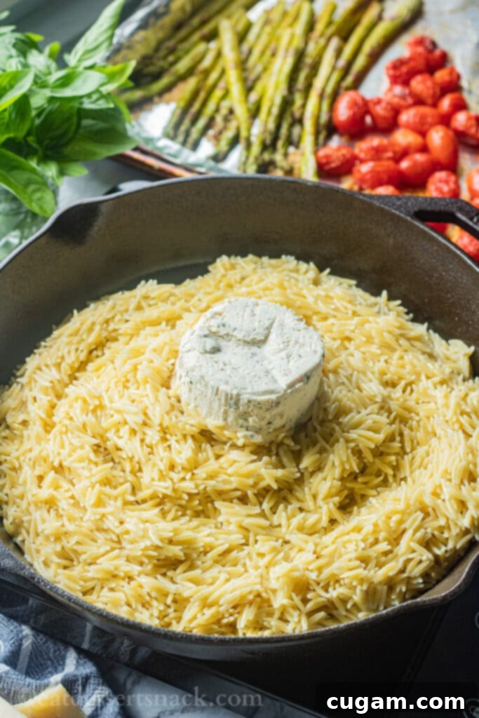 Cooked orzo pasta in a pan with a block of Boursin cheese placed on top, ready to be stirred in.