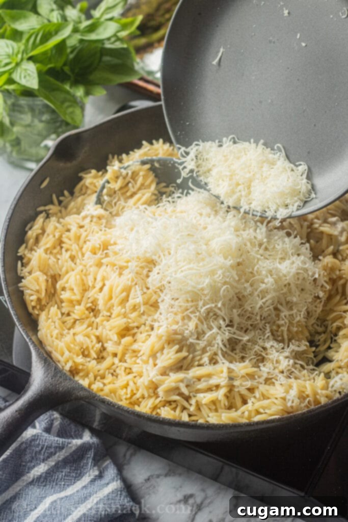 Pouring a large amount of freshly grated Parmesan cheese onto a pan of cooked creamy orzo pasta.