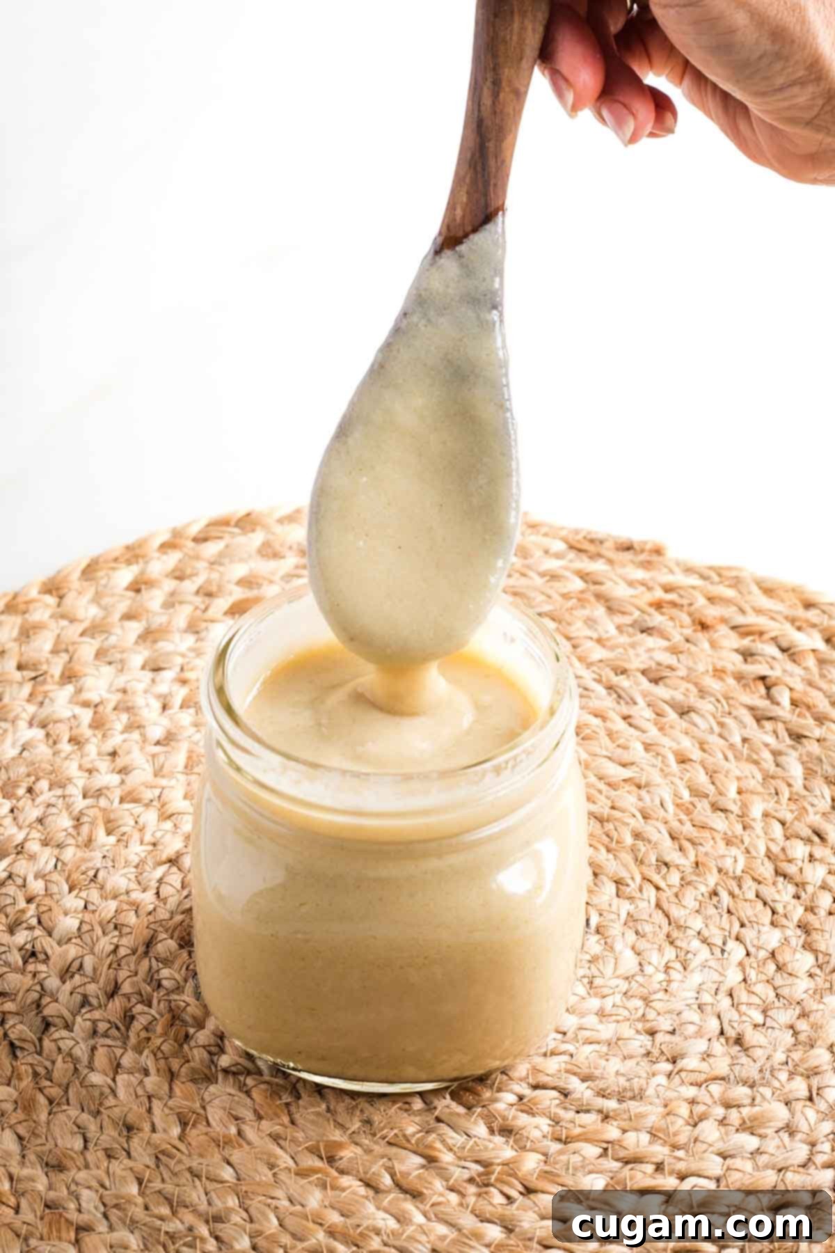 A wooden spoon, coated with thick, creamy vegan bechamel sauce, emerging from a glass jar, demonstrating the sauce's perfect consistency.