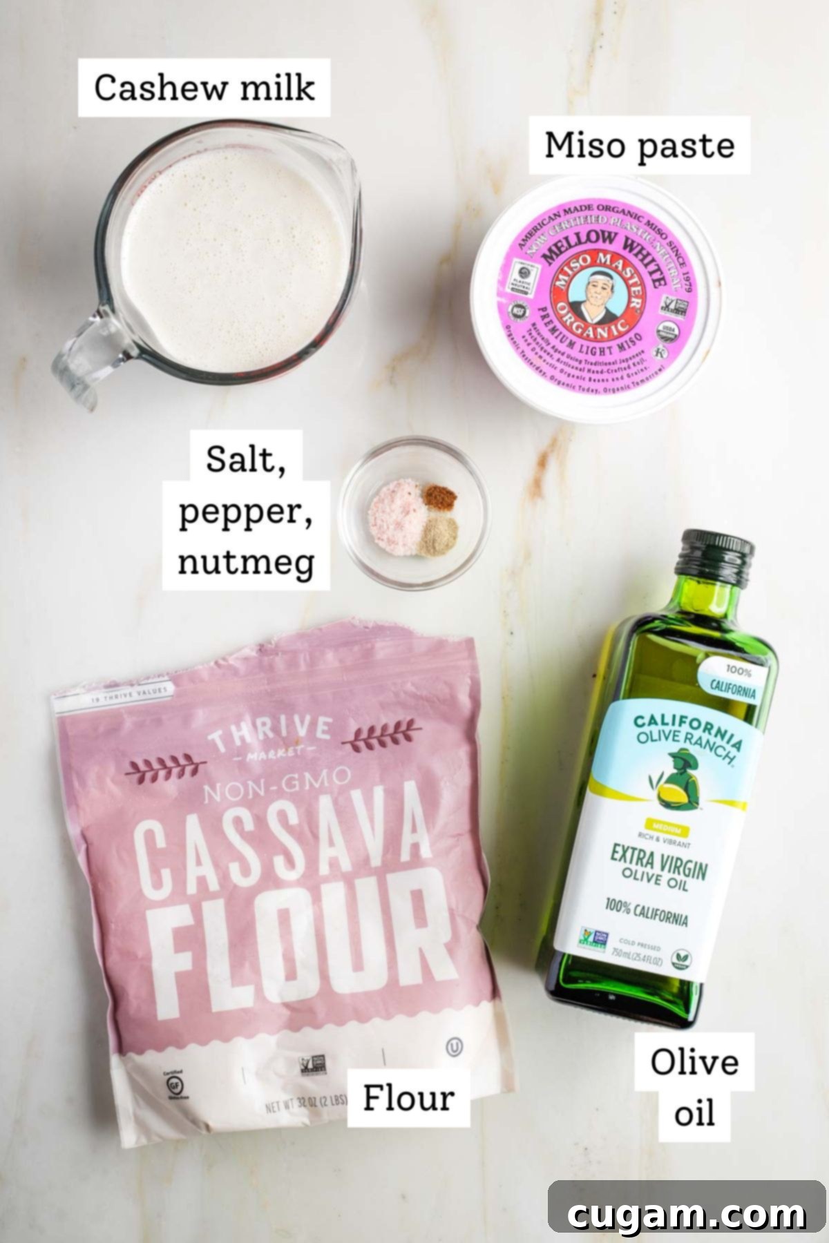 A selection of ingredients for vegan bechamel sauce laid out on a wooden surface, including a bowl of cashew milk, olive oil, cassava flour, a spoon of miso paste, and spices.