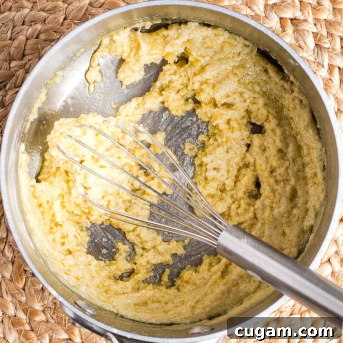 A whisk vigorously stirring a super thick, pasty, pale yellow mixture (the roux) in a small saucepan, showing its consistency.