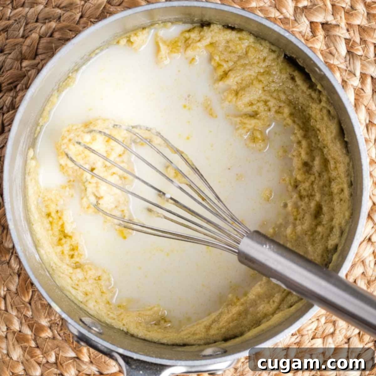 A whisk stirring plant-based milk into a thick, light yellow, pasty mixture in a saucepan, beginning to loosen the roux.