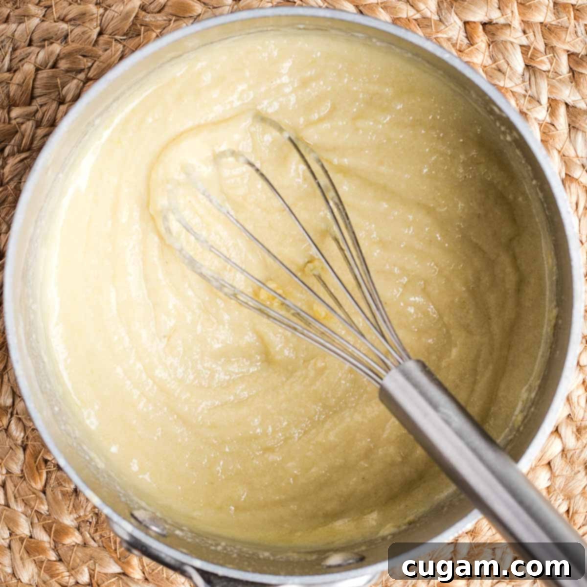 A whisk actively stirring a pale yellow, slightly thickened vegan bechamel sauce in a small pot, demonstrating the emulsification process.