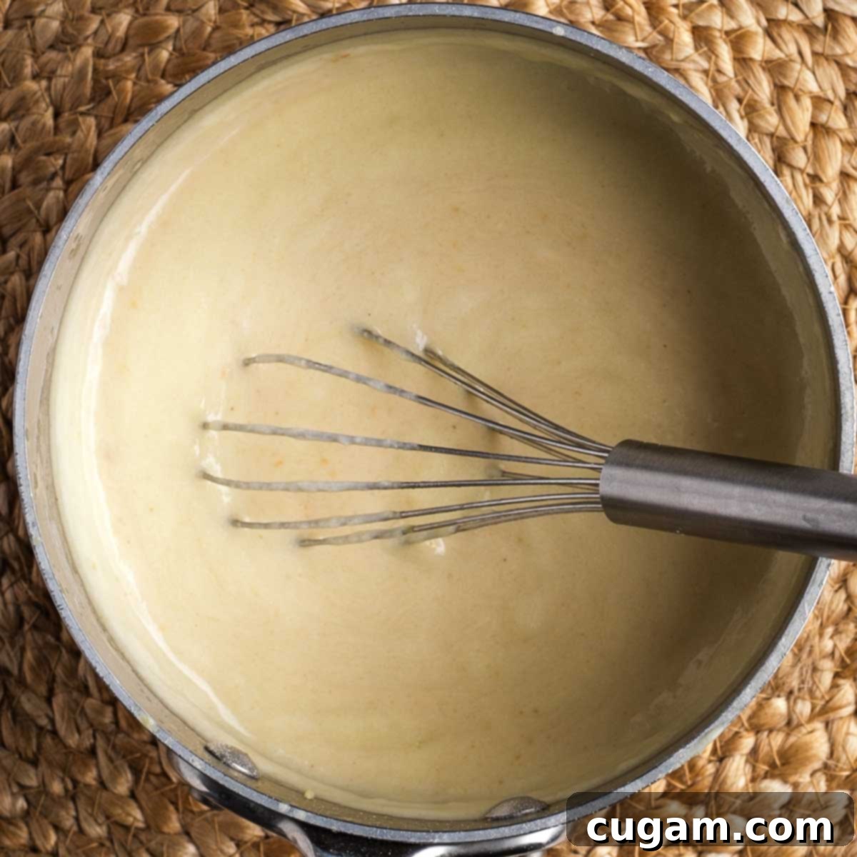 A whisk stirring a thick, beige-colored vegan bechamel sauce in a small saucepan, indicating its ready consistency.