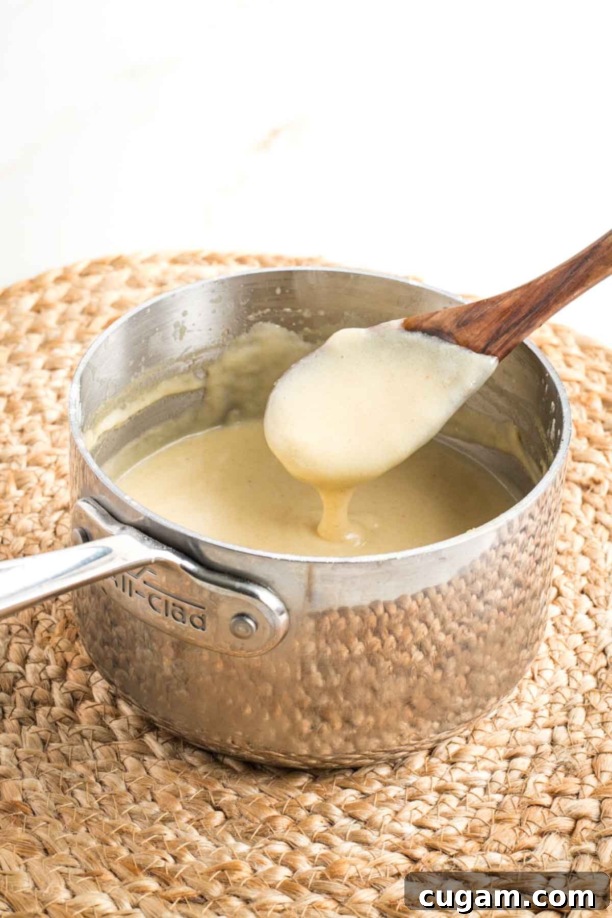 A wooden spoon peeking out of a saucepan full of creamy white vegan bechamel sauce to show how thick and perfectly smooth it is, ready for serving.