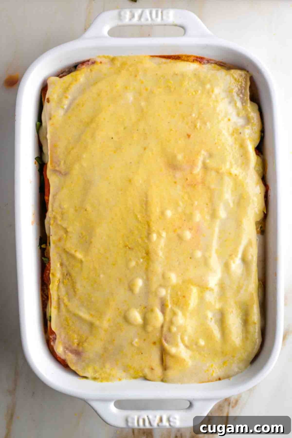 Casserole dish layered with sheets of lasagna noodles, red sauce, white sauce, zucchini and greens, ending with bechamel and nutritional yeast.