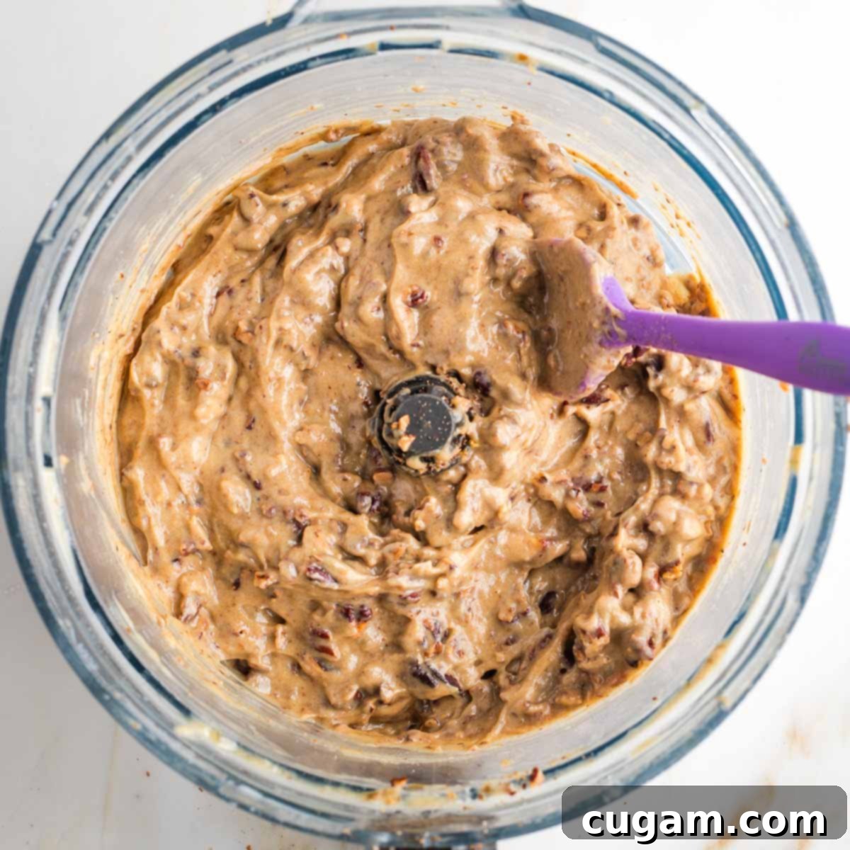 Golden colored caramel spread with chopped pecans in the bowl of a food processor with silicon spatula stirring. 