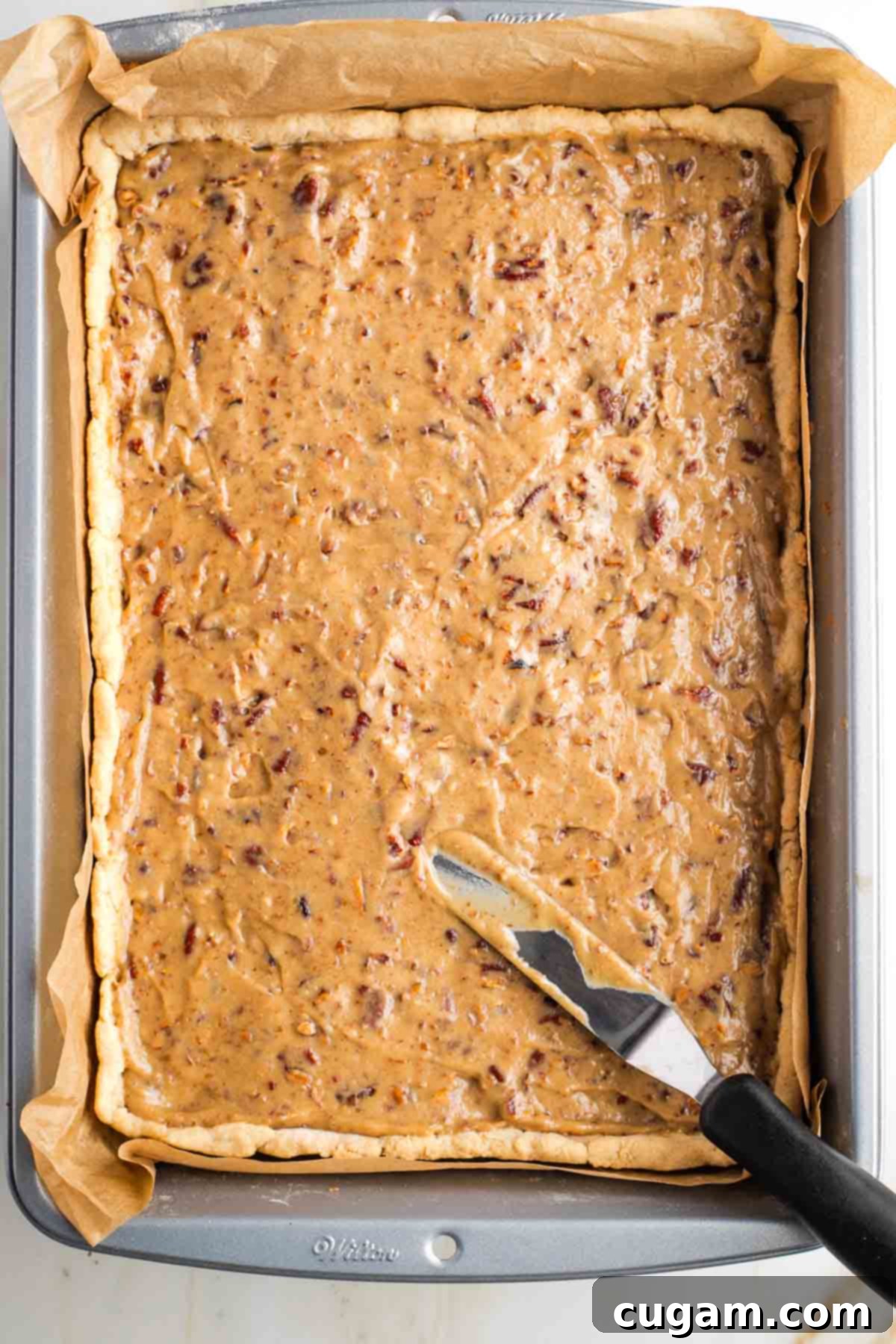 Offset spatula spreading caramel pecan spread onto a pie crust in a rectangular baking pan. 