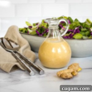 Effortless Ginger Miso Tahini Drizzle 11 Glass pitcher of ginger miso dressing with a fresh green salad and ginger root in the background, ready to be served.