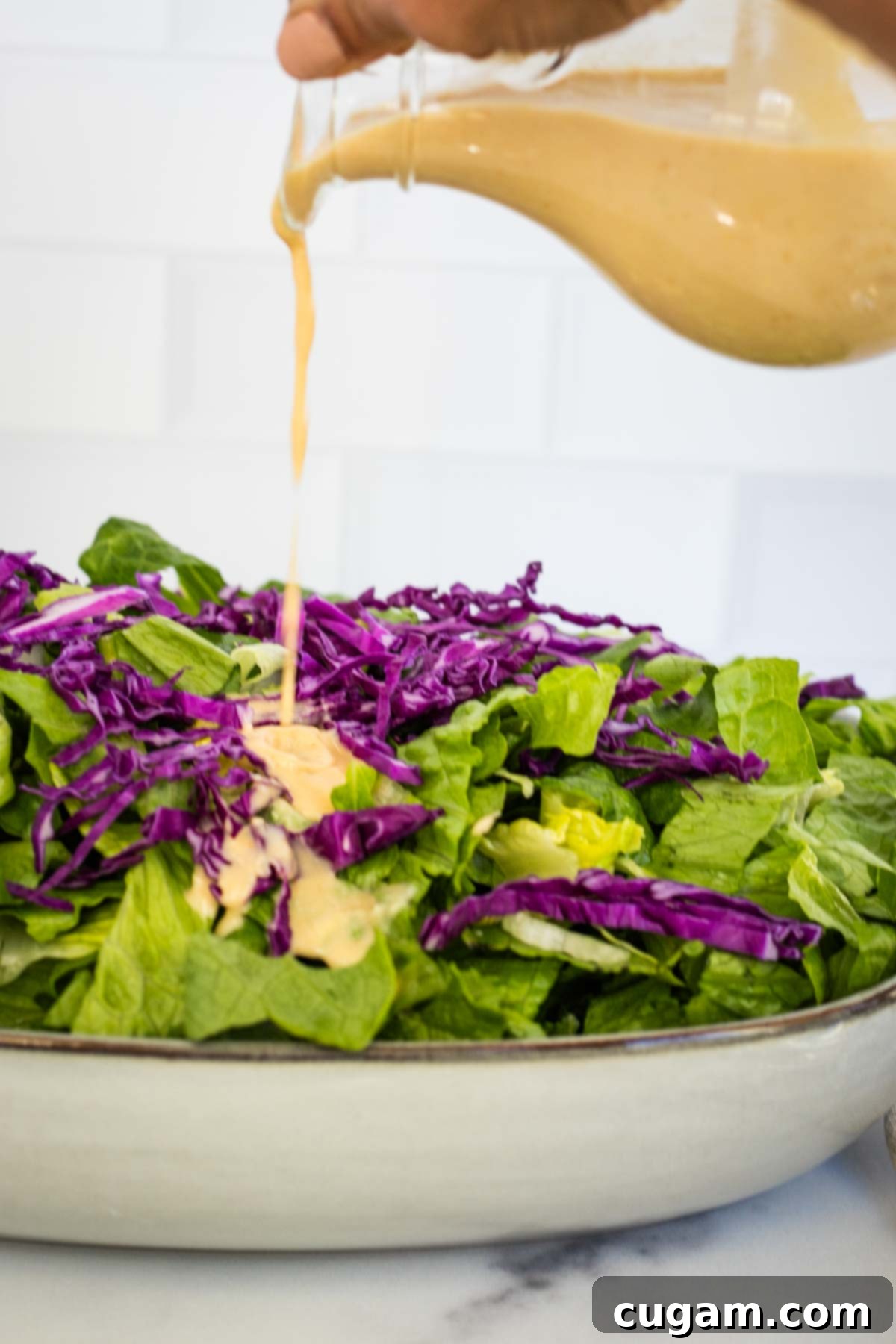 Effortless Ginger Miso Tahini Drizzle 8 Pouring ginger miso dressing out of a glass pitcher onto a mixed green salad, featuring vibrant greens and purple cabbage.