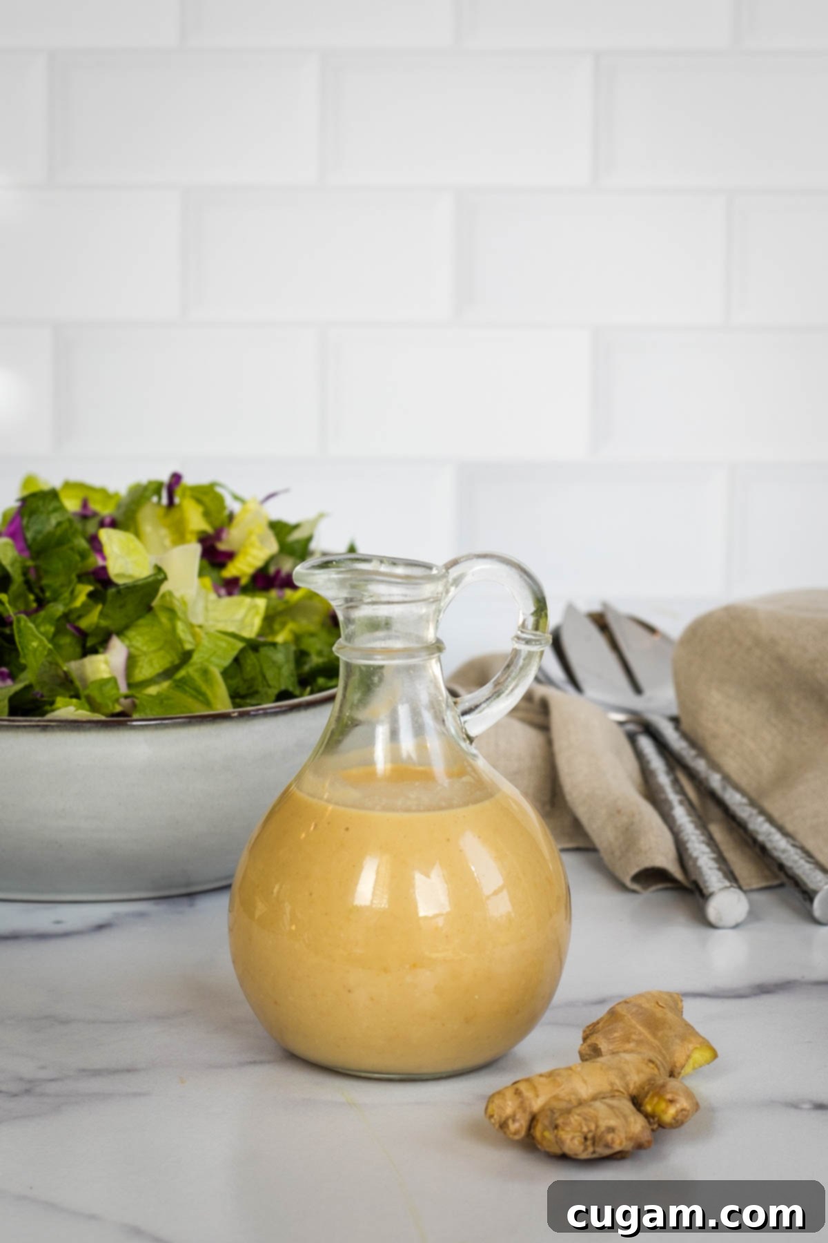 Effortless Ginger Miso Tahini Drizzle 10 Small glass pitcher of creamy miso dressing with a knob of fresh ginger in front and a bowl of mixed salad in the background, showcasing the dressing's uses.