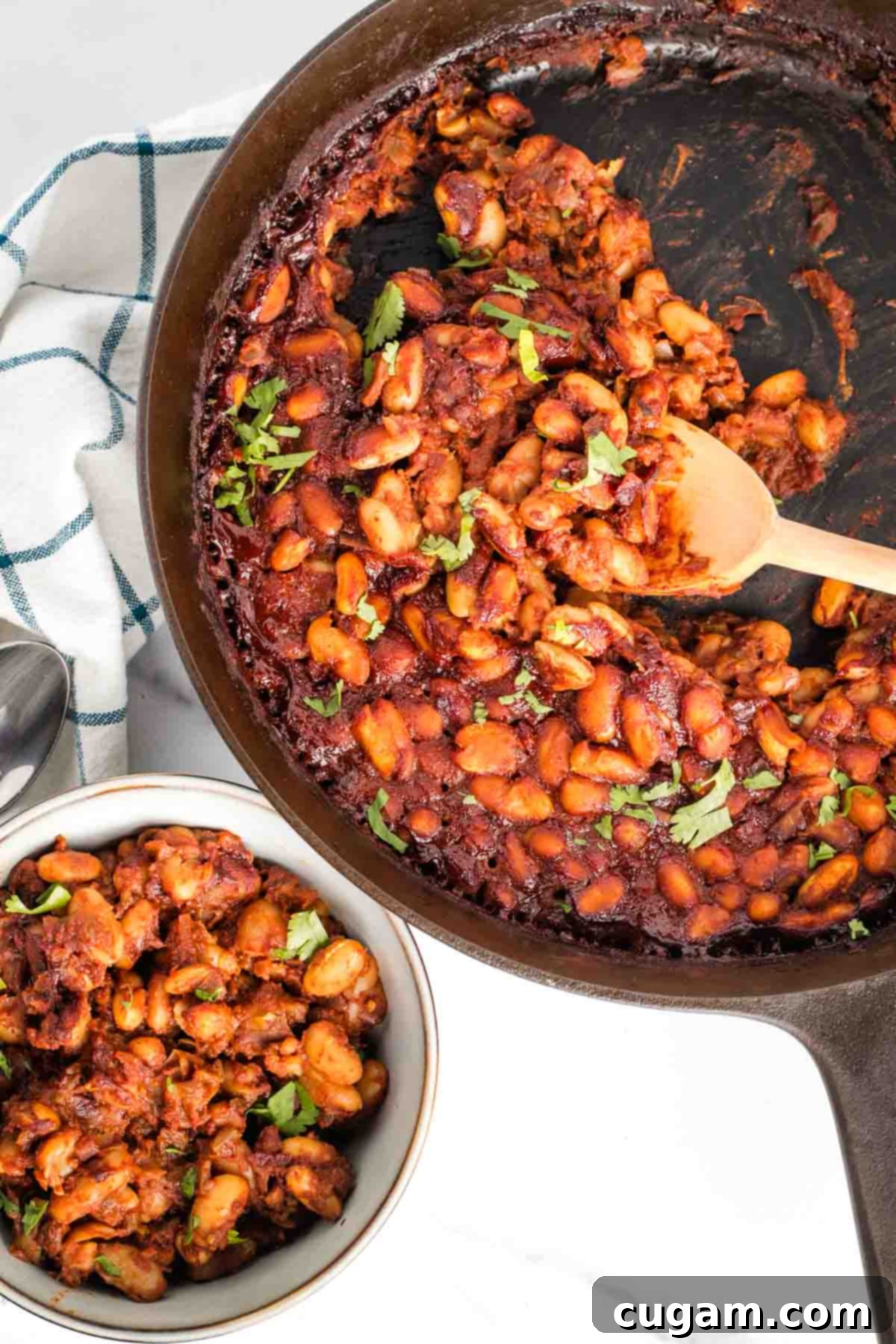 Skillet of delicious vegan BBQ baked beans, generously sprinkled with fresh herbs, with a wooden spoon scooping some into a small bowl, ready to be served.