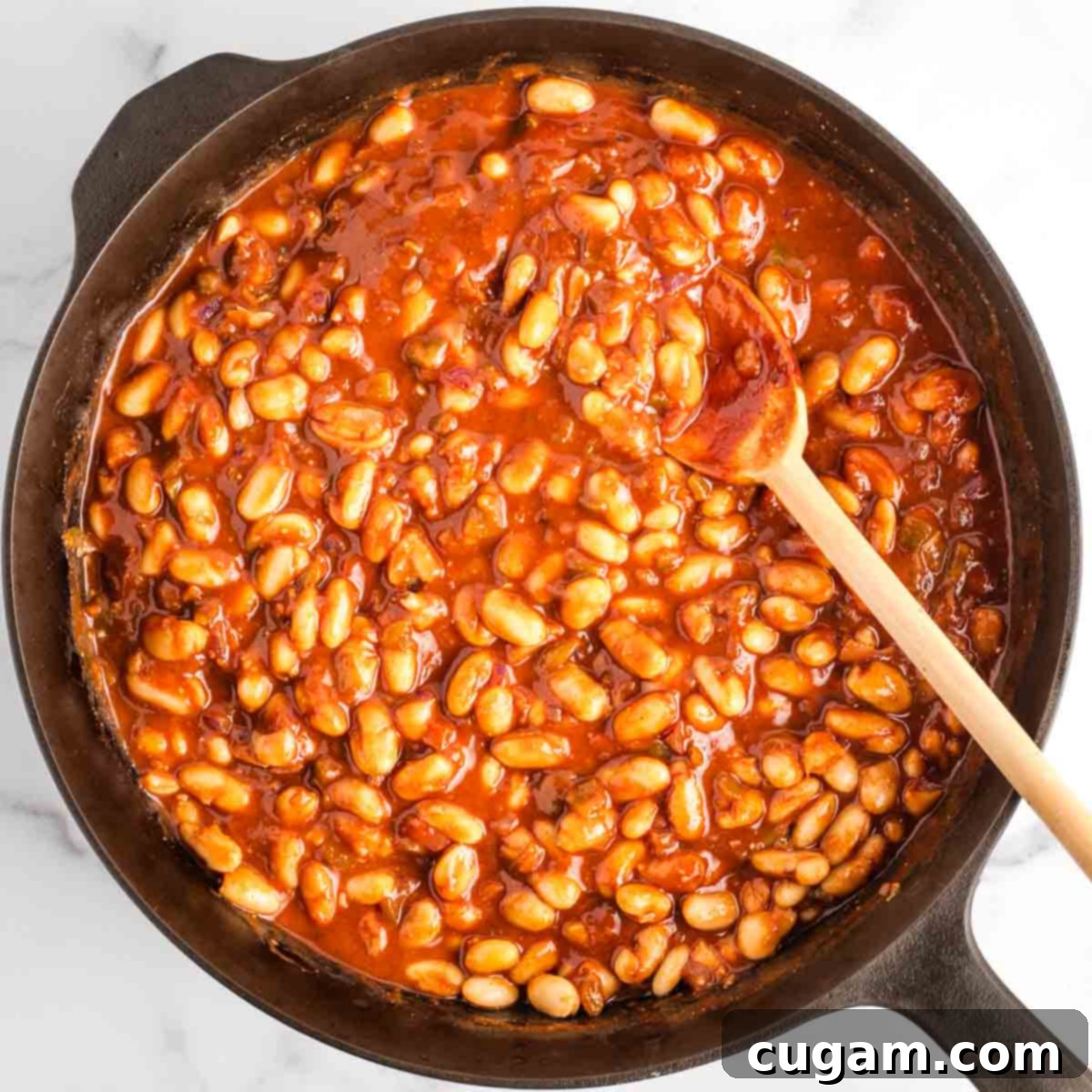 A wooden spoon stirring cannellini beans generously coated in a rich, thick, tomato-based BBQ sauce within a cast iron skillet, ensuring all ingredients are well combined.