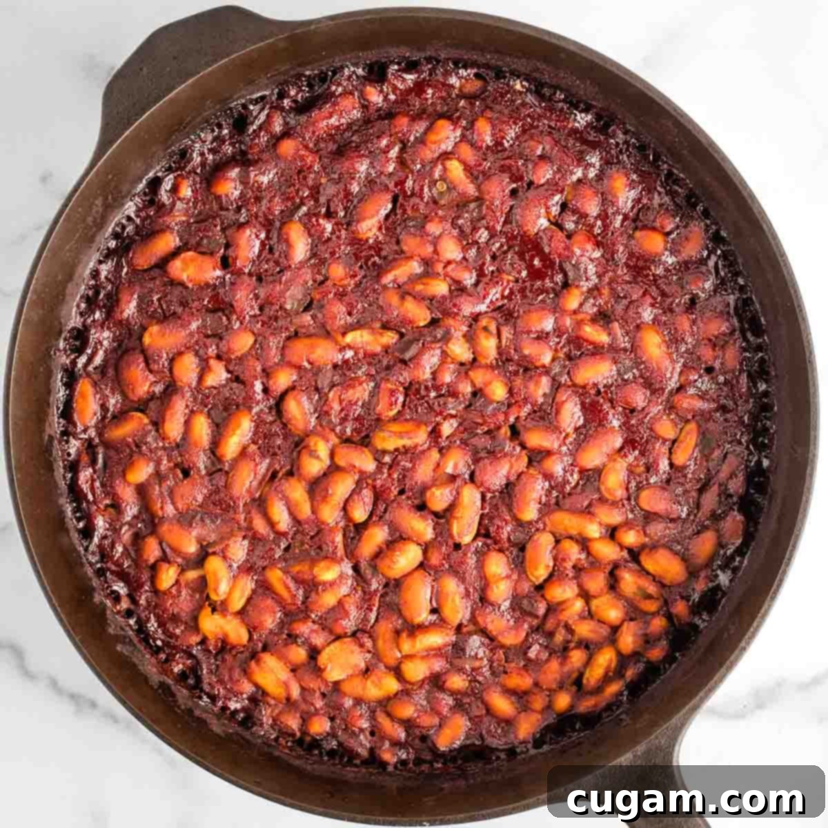 A cast iron skillet filled with bubbling, deep red BBQ baked beans, just out of the oven, with a rich, glossy sauce and perfectly tender beans.