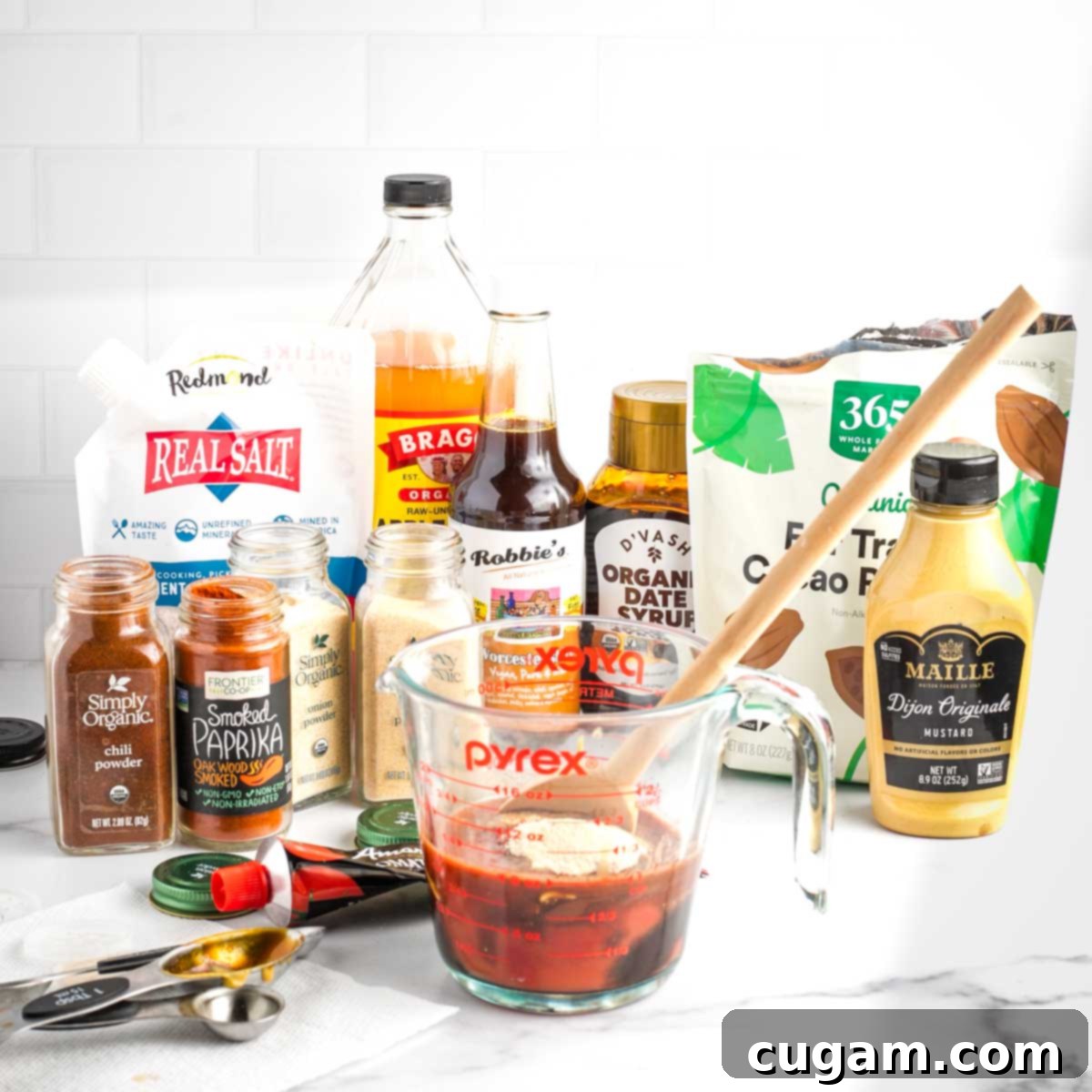 An assortment of bottles, jars, and spices, with a Pyrex measuring cup in the foreground, ready for stirring up a batch of easy homemade BBQ sauce.