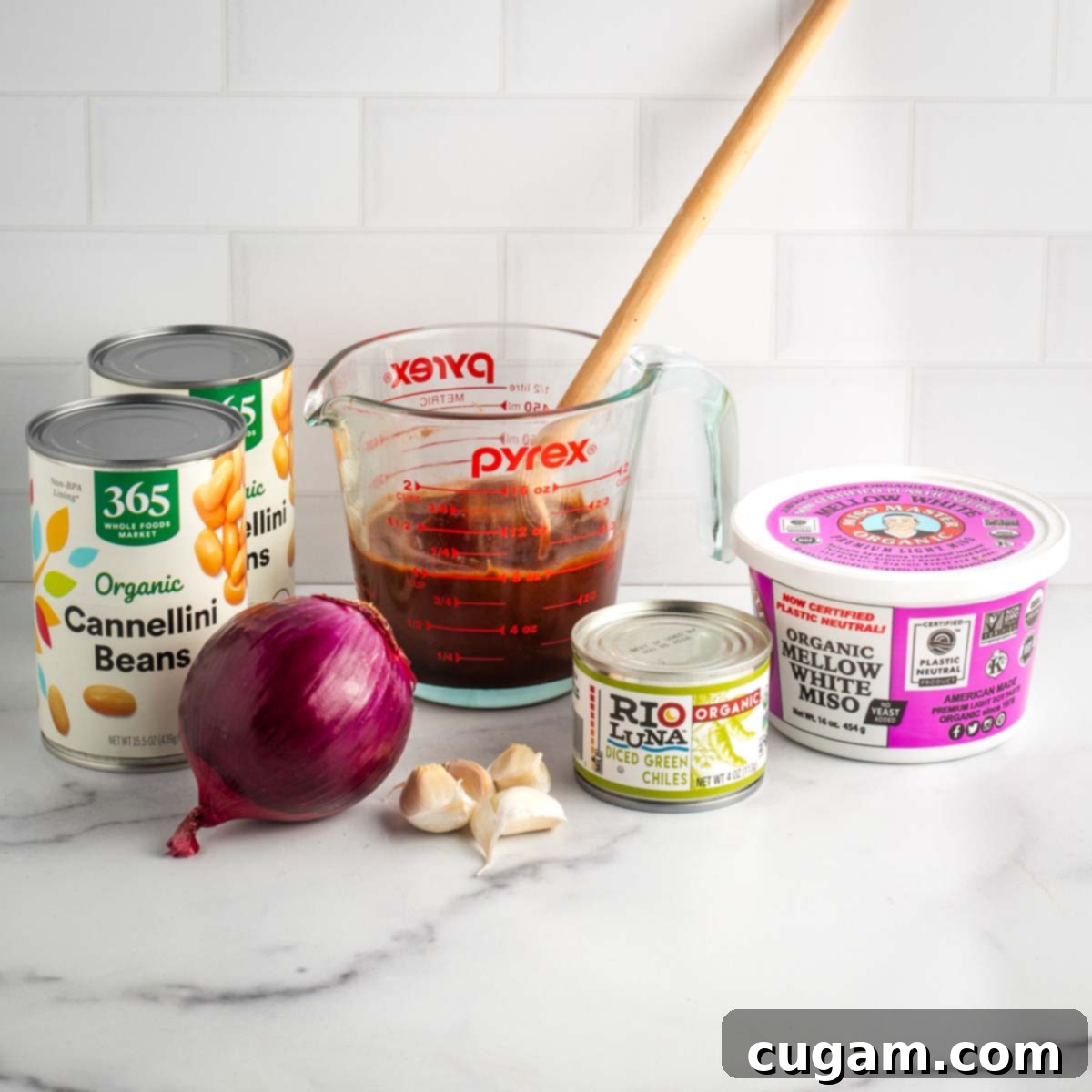 A collection of ingredients for vegan baked beans: a measuring cup of homemade BBQ sauce, cans of cannellini beans, a red onion, garlic cloves, a can of diced green chiles, and a container of miso paste, all laid out on a kitchen counter.