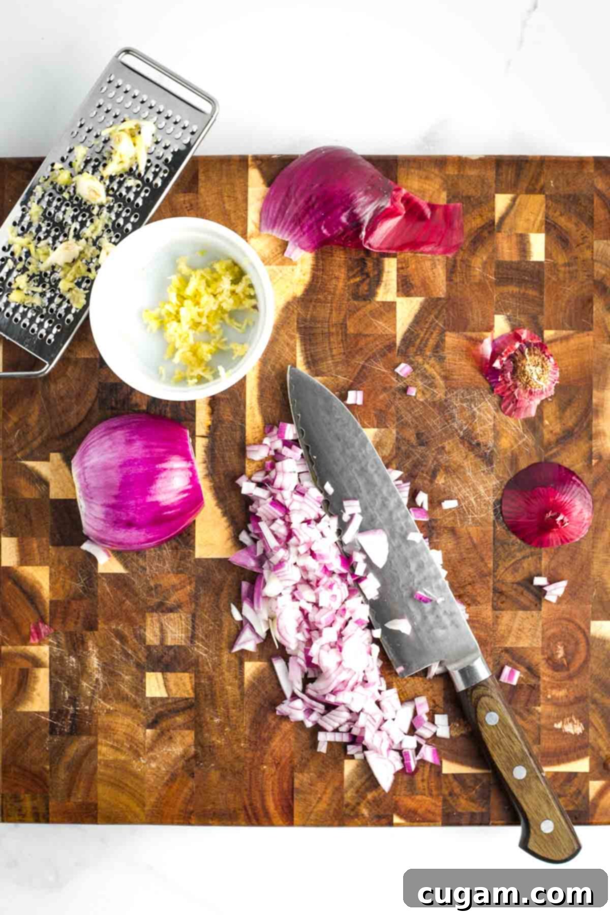 A wooden cutting board with a red onion being diced and a small dish holding finely grated garlic cloves, illustrating the prep work for the baked beans recipe.