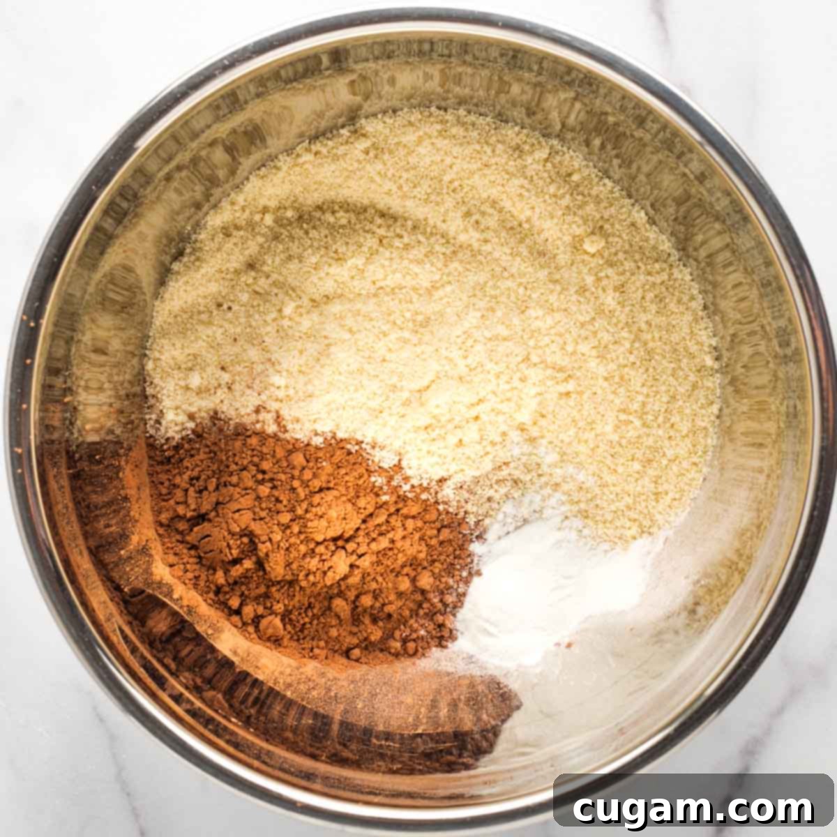 Metal mixing bowl containing almond flour, cacao powder, and baking powder, perfectly blended with a fork.