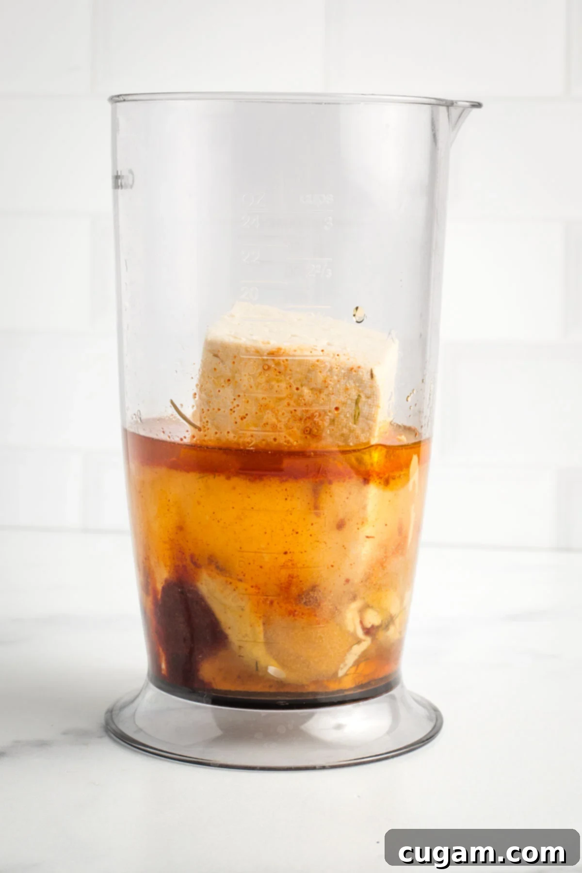 Tall container with a block of extra firm tofu, along with measured pickle juice, water, maple syrup, spices, and mustard.
