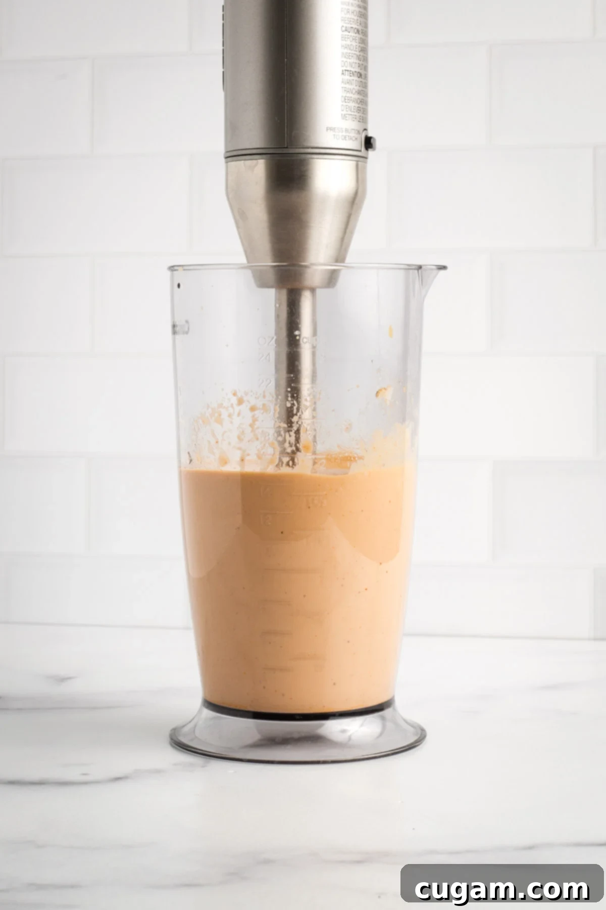 An immersion blender pureeing the peach-colored sauce in a tall container until smooth and uniform.