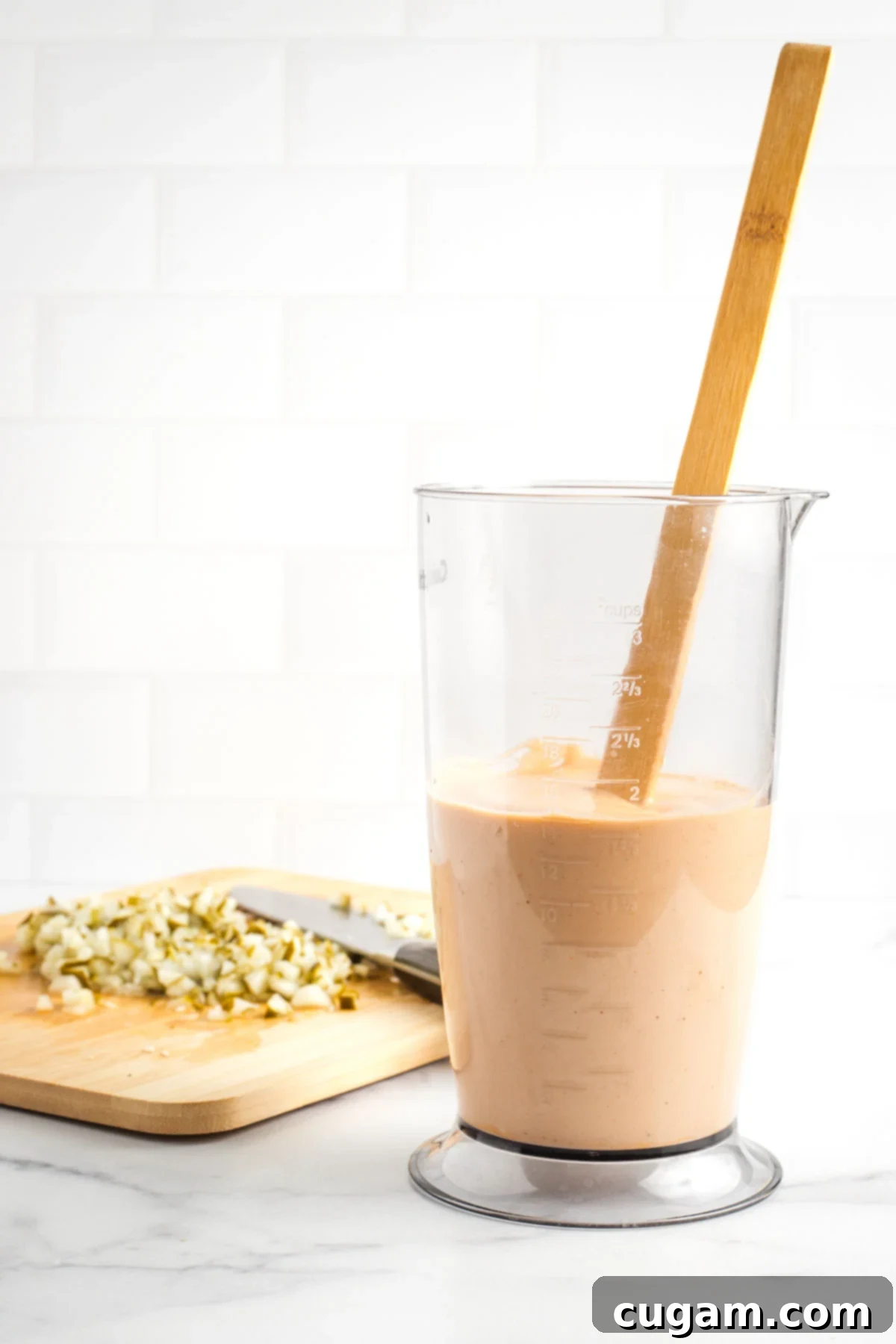 The smooth, peach-colored burger sauce in a tall container, with finely diced pickles ready on a board in the background.