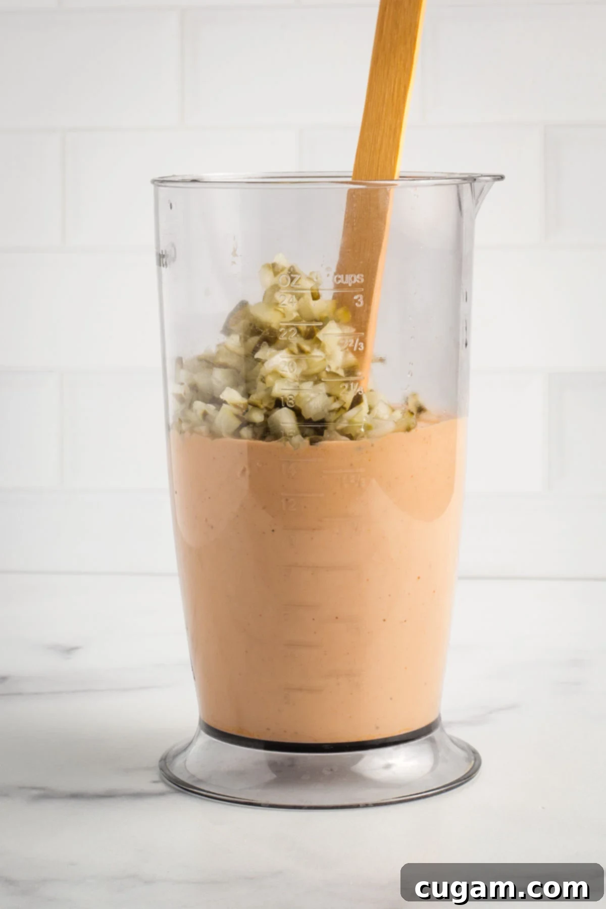 Adding the finely diced pickles directly into the tall container of creamy burger sauce.