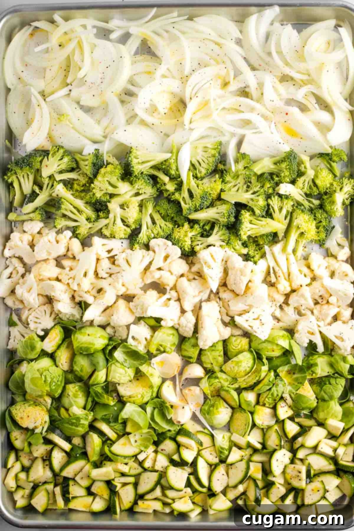 Rows of chopped veggies on a sheet pan. Onion, zucchini, cauliflower, brussels sprouts and some cloves of garlic with the skins still on.