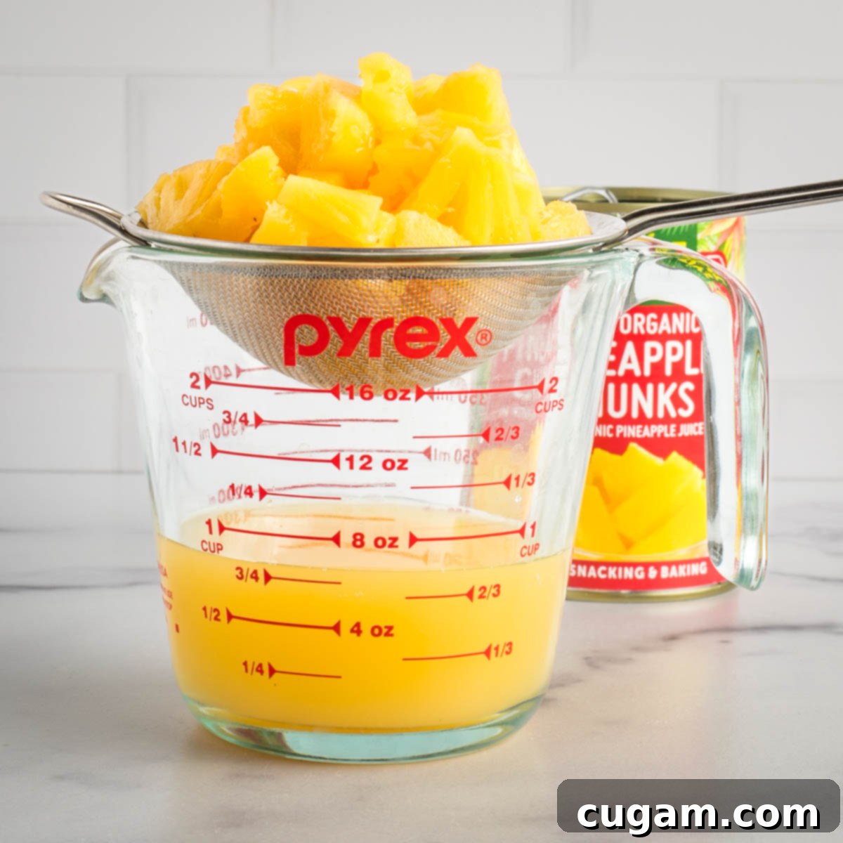 Strainer, set over a glass measuring cup separating canned pineapple chunks from the juice. 