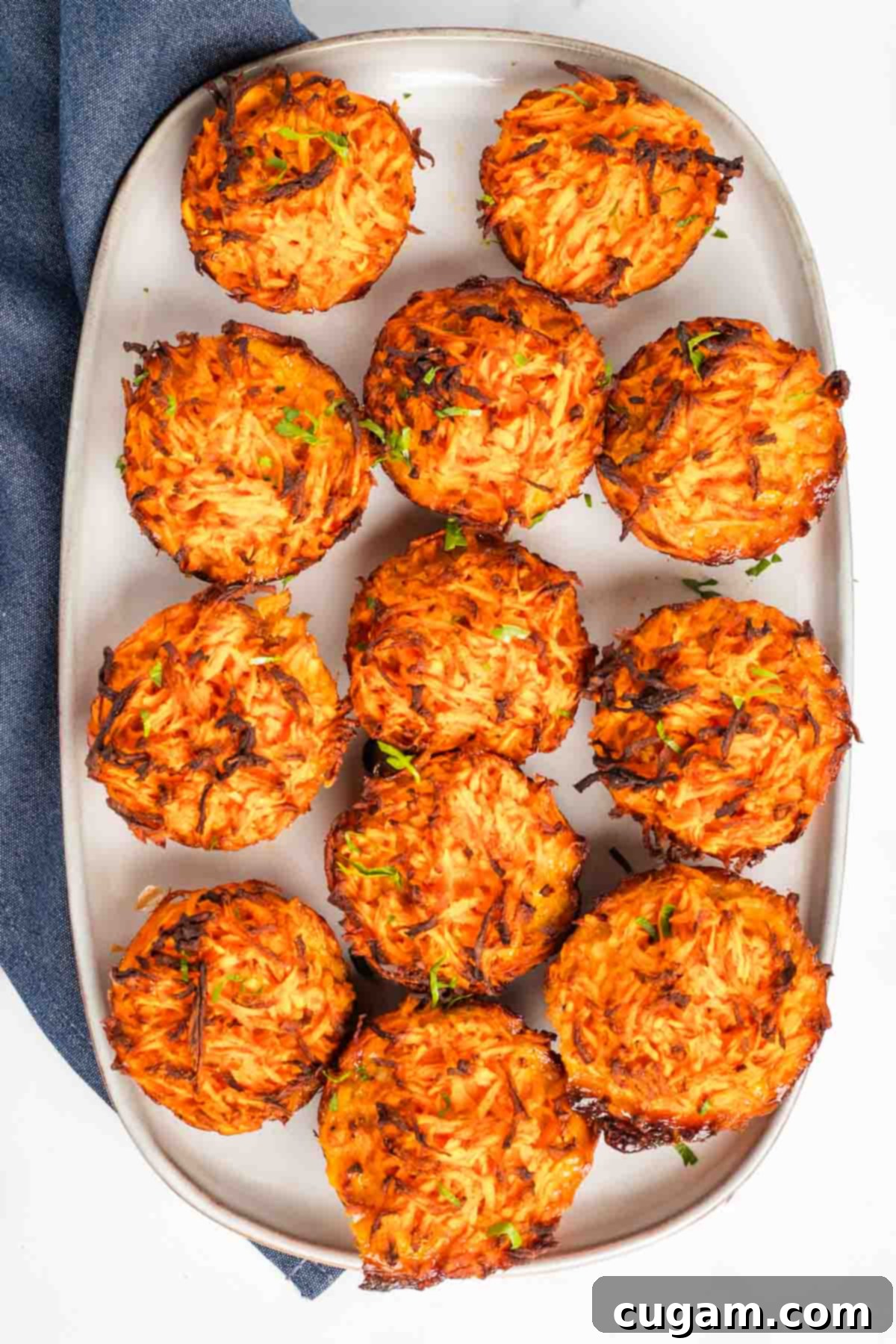 Crispy sweet potato kugel muffins on a plate, sprinkled with fresh herbs. Each kugel is perfectly browned with a golden-orange hue, making them look incredibly appetizing. They are arranged neatly, suggesting a perfect dish for entertaining or a healthy meal.