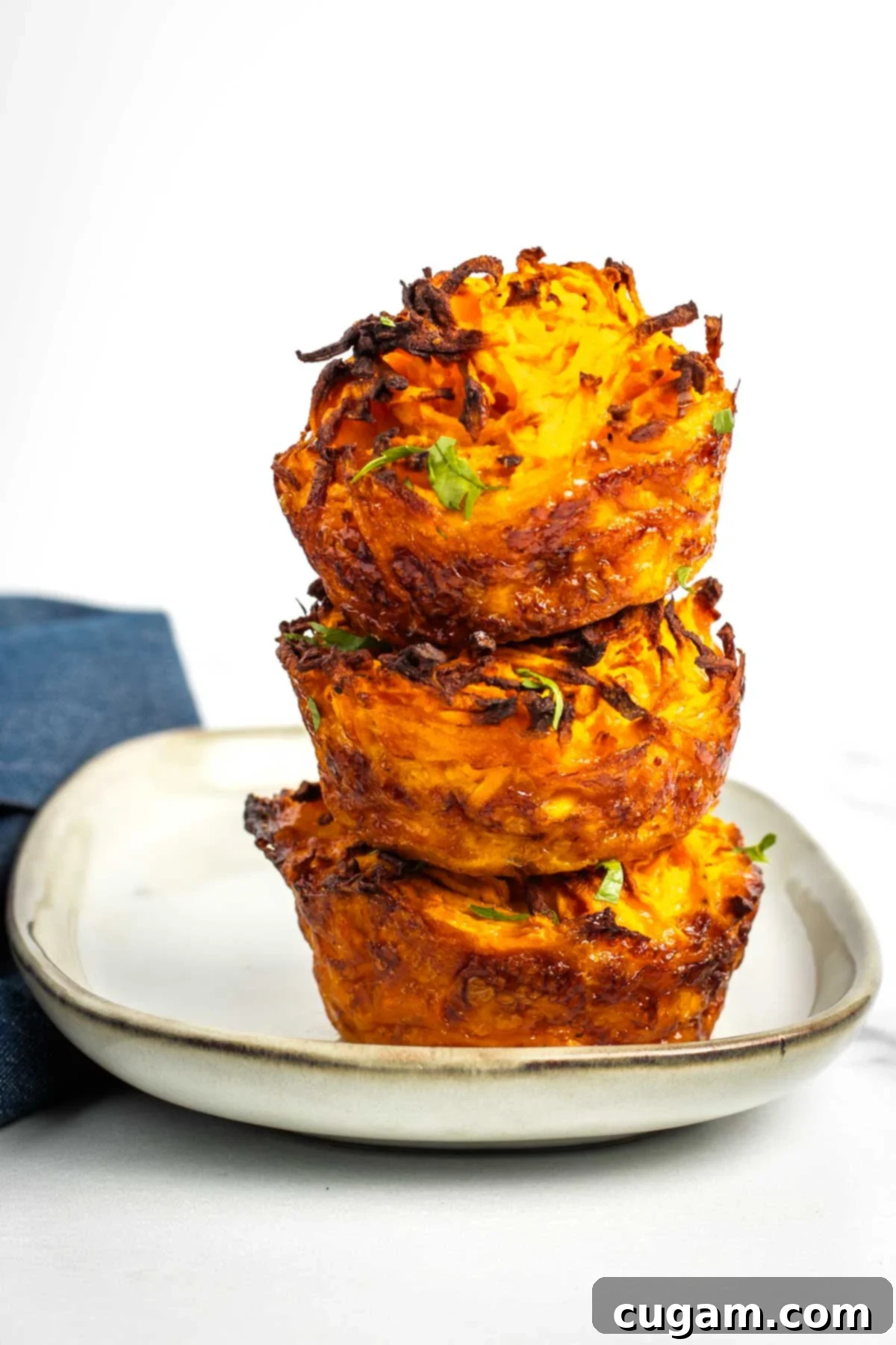 Close-up of a single crispy sweet potato kugel cup, garnished with fresh herbs, on a white plate. The kugel shows off its golden-brown, textured exterior and hints of tender inside.